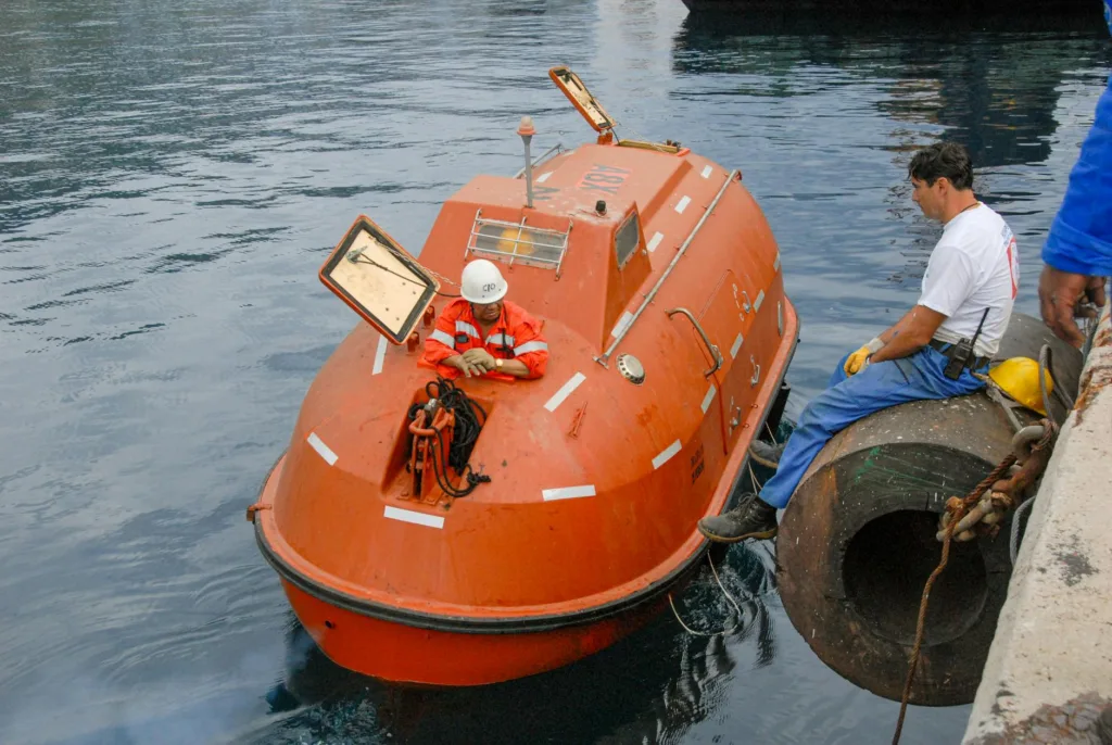 Latihan dan Prosedur Darurat 6 Crew members prepare a lifeboat for launch at a harbor. Maritime safety equipment and procedures.